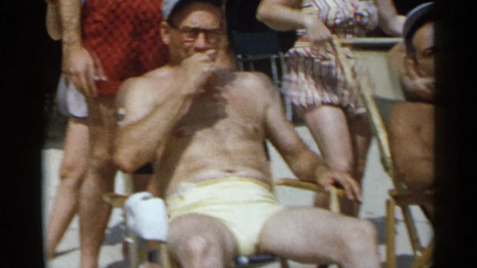 1954 New York beach home-movie frame showing a seated man in yellow trunks with adults standing nearby.