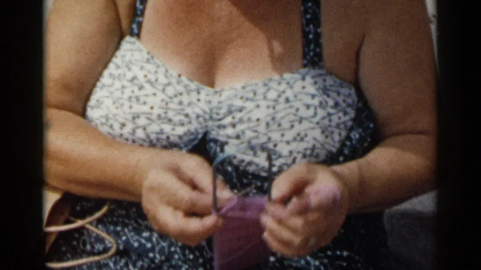1954 New York home-movie frame showing a seated woman in a patterned swimsuit knitting purple yarn near her lap.