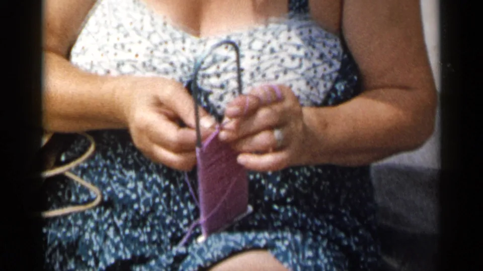 Close 1954 New York home-movie frame of a woman using a small metal loom with purple yarn while seated in a swimsuit.