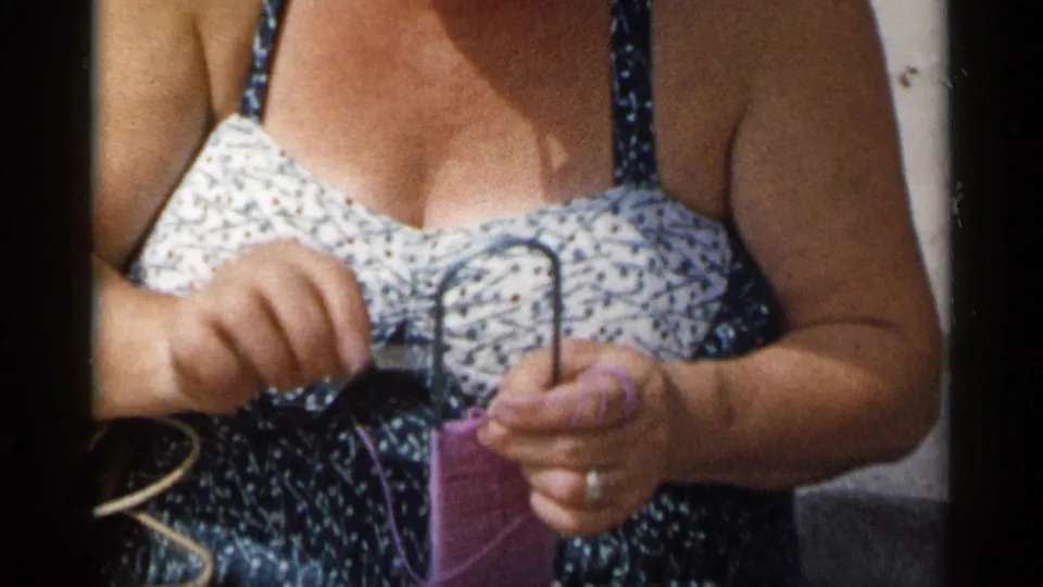 In supported 1954 New York home-movie footage, a woman in a floral bathing suit begins knitting with purple yarn on a sunny beach.