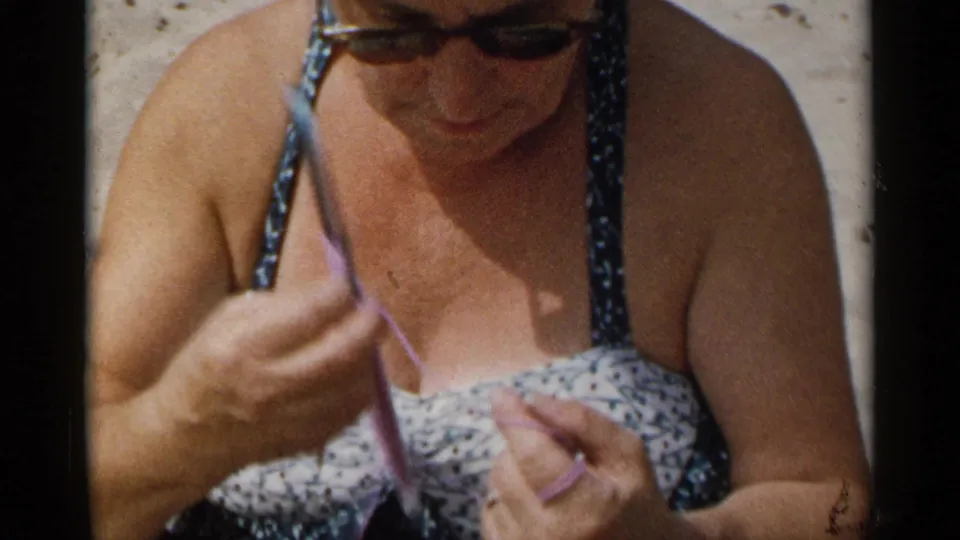 A woman in sunglasses and a floral bathing suit looks down at purple yarn and knitting needles in the 1954 beach footage.