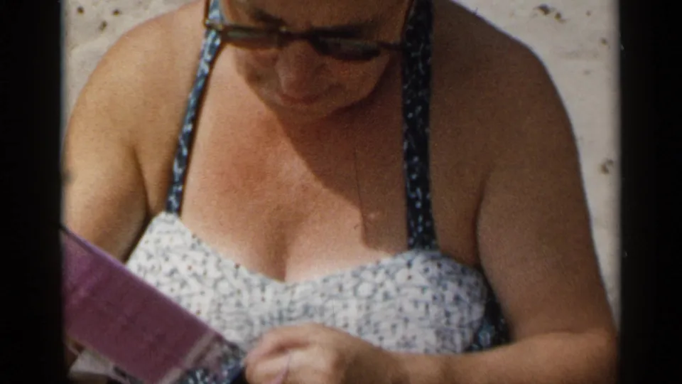 The seated woman remains focused on a purple knitted piece, with her floral bathing suit and sunglasses visible in the beach scene.