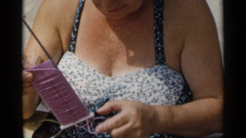 A close view shows the woman holding a purple knitted panel and needle while seated outdoors in the supported 1954 New York beach footage.