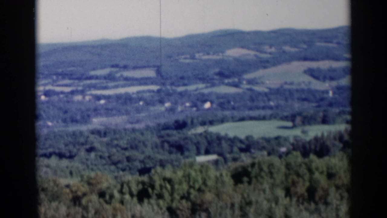 1955 Lake George 8mm view of tree-covered hills, open fields, and distant scattered buildings beneath a pale sky.