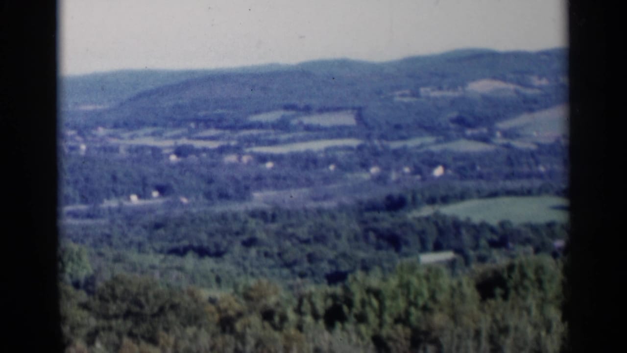 Lake George landscape with layered wooded hills, patchwork fields, and small distant buildings in soft 8mm color.