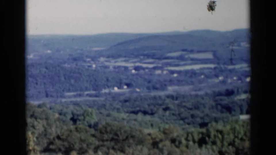 1955 home-movie frame showing a hazy Lake George area valley with wooded slopes, fields, and scattered buildings.