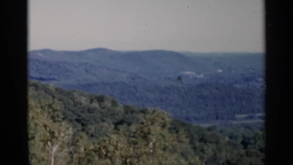 1955 Lake George 8mm frame showing a broad green hillside, distant valley openings, and blue ridges beyond.