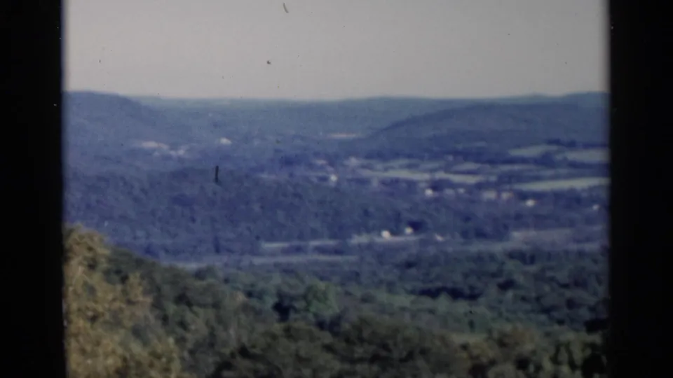 1955 Lake George-area 8mm frame showing wooded foreground, rolling hills, scattered fields, and distant ridges.