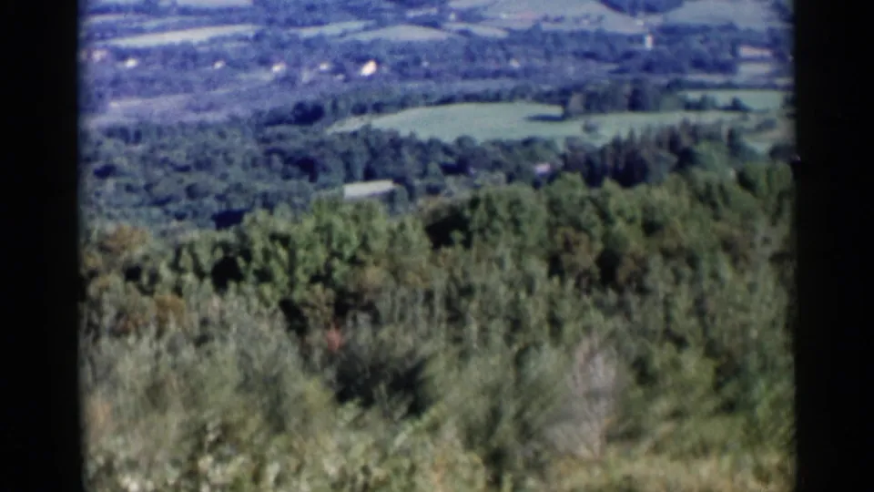 1955 Lake George-area 8mm still showing a wooded foreground, valley fields, scattered structures, and hazy hills beyond.