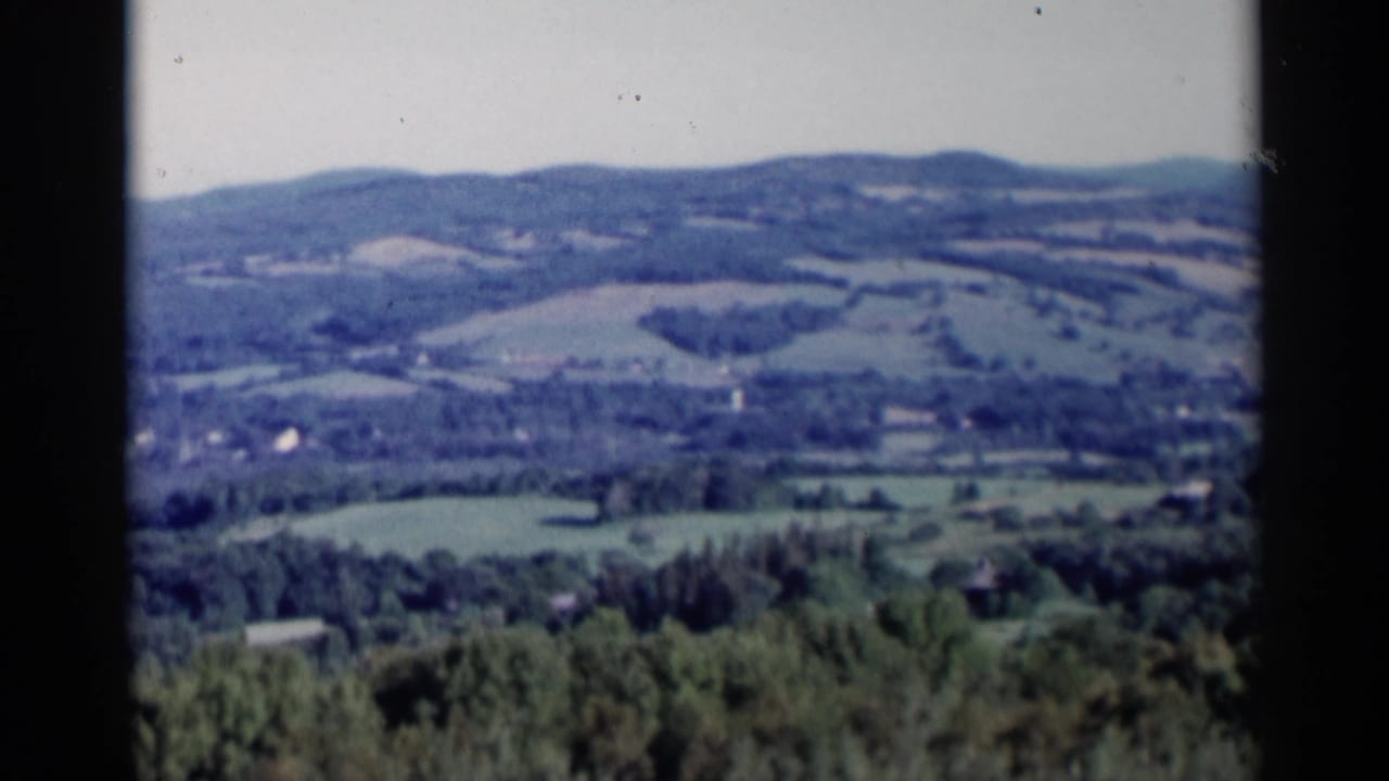 Panoramic countryside view with tree-covered slopes, fields, and distant hills.