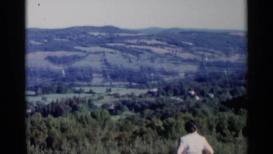 1955 Lake George home-movie frame with a woman in a white shirt at the overlook below wooded hills and distant fields.