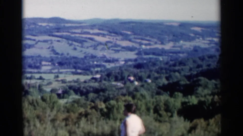 Lake George area 8mm frame showing a woman from behind at a scenic overlook, facing wooded hills and open countryside.