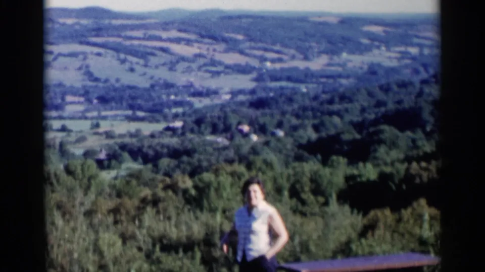 1955 Lake George home-movie frame with a woman near the foreground before a broad wooded valley and hazy hills.