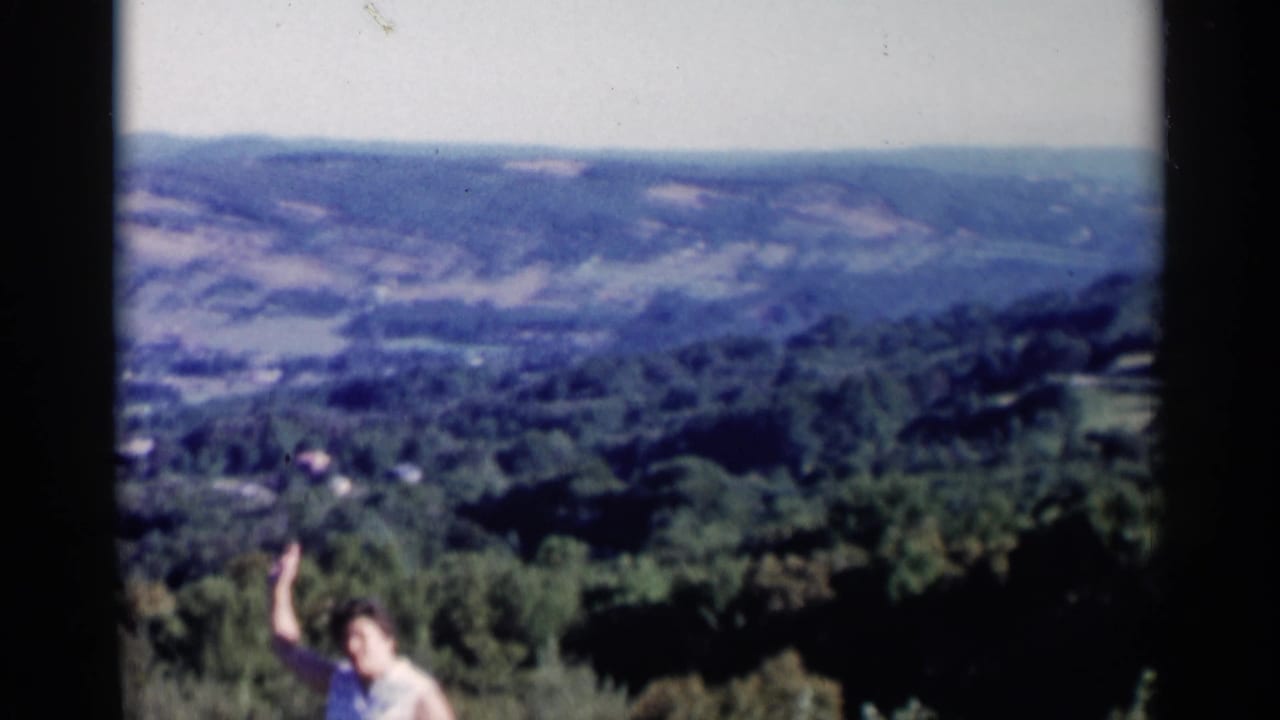 1955 Lake George frame showing a woman raising her arm toward the camera against wooded hills and a distant valley.