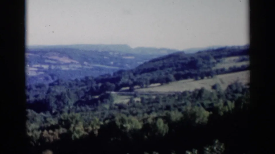 1955 Lake George frame showing a quiet wooded valley, sloped fields, and hazy hills from an elevated viewpoint.