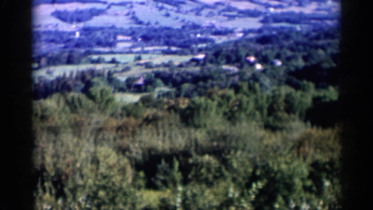 Lake George, New York countryside appears in a soft 8mm frame with wooded slopes, fields, and distant houses.