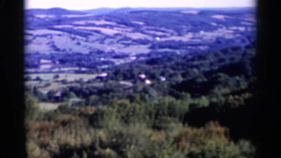 Wide 1955 Lake George countryside frame with hills, wooded slopes, fields, and small houses in the distance.