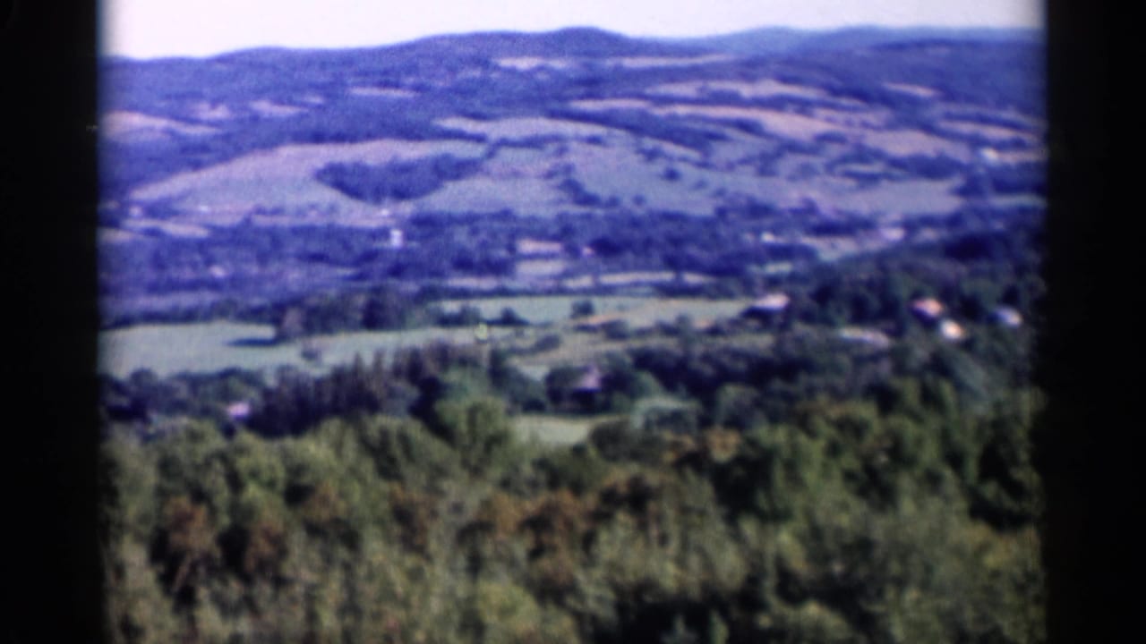 1955 Lake George countryside view with blue-tinted 8mm color, wooded slopes, open fields, and a few distant structures.