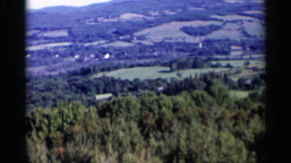 1955 Lake George 8mm frame looking across a wooded foreground toward distant fields, hills, and small rural structures.