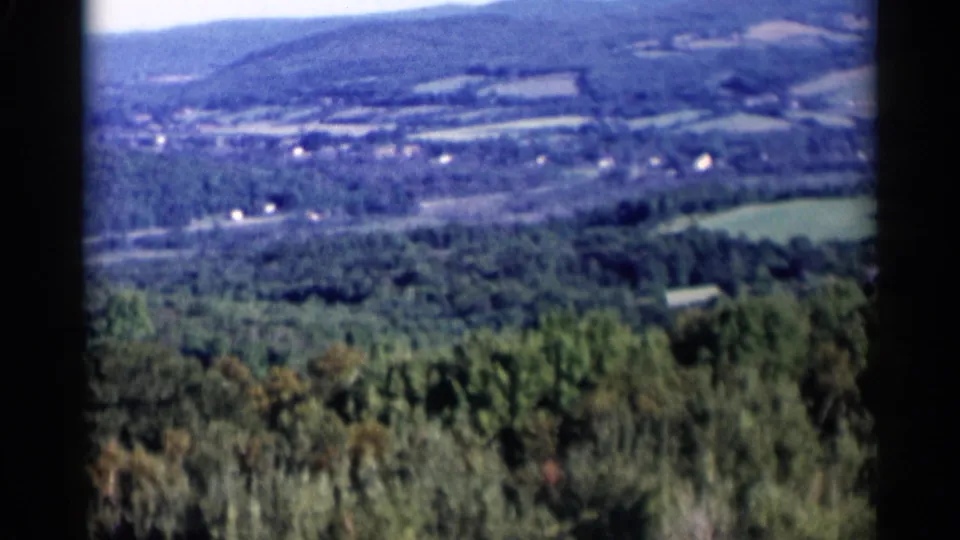 1955 Lake George home-movie frame showing treetops below, fields across the valley, and hazy wooded hills in the distance.