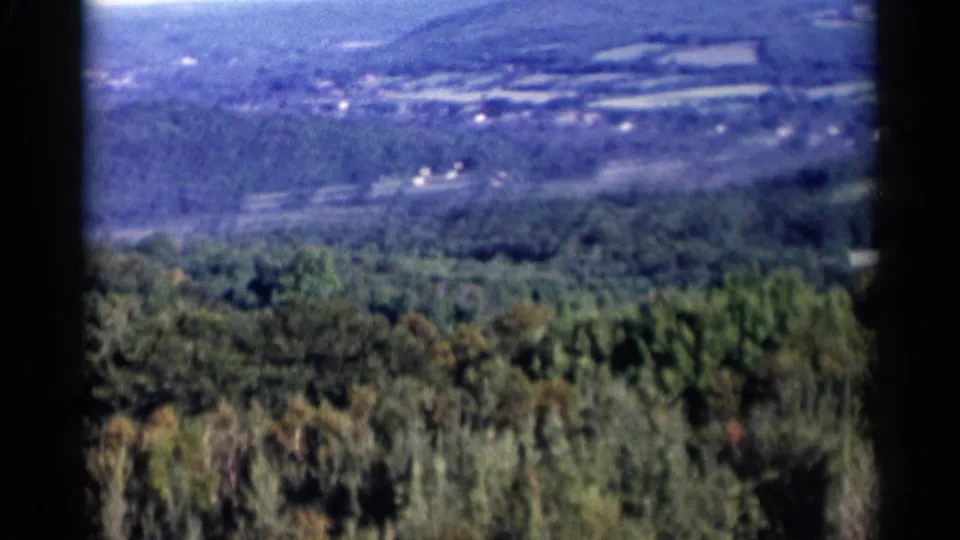 1955 Lake George-area 8mm frame showing a soft aerial view of forested hills with fields and scattered buildings in the distance.