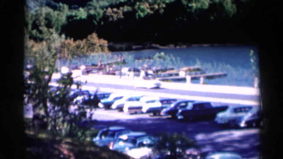 Lake George lakefront view with mid-century cars parked beside a road, docks and boats near the water.