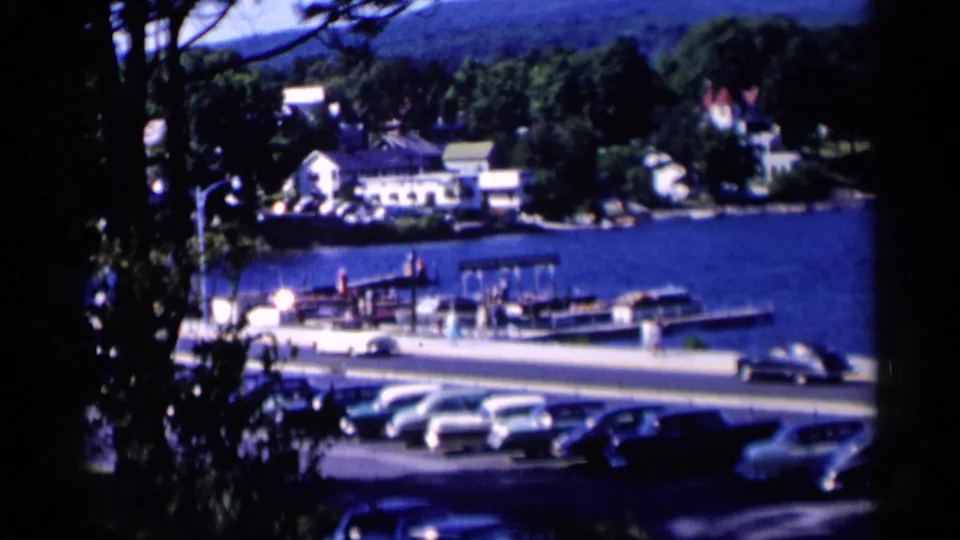 Lake George, New York view framed by trees, showing parked cars, marina docks, boats, and shoreline buildings.