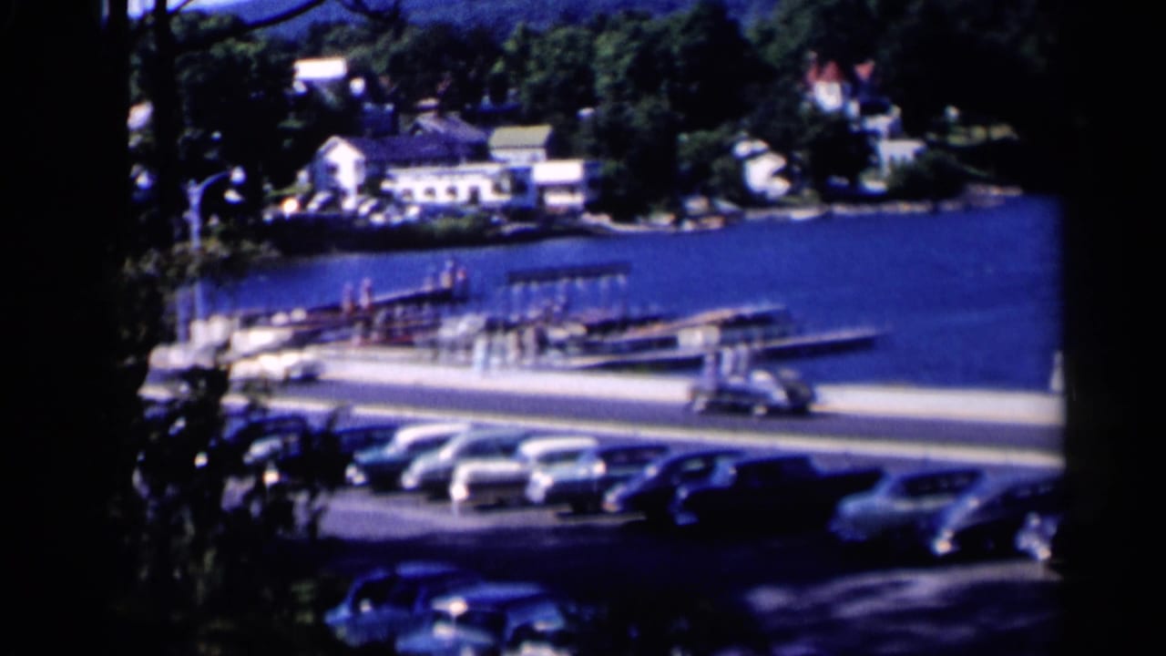 Lakeside road with parked cars, docks, boats, trees, and houses across the water.