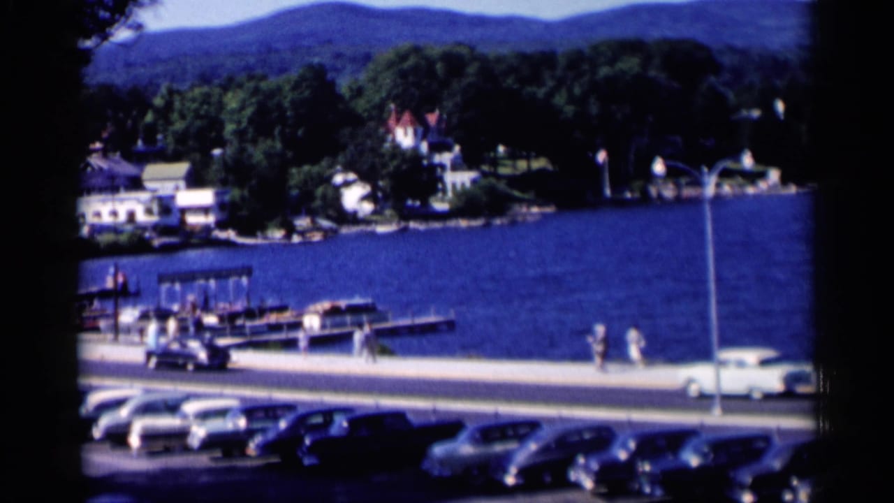 Pedestrians walk near a lakefront road with parked cars, docks, boats, and houses across the water.
