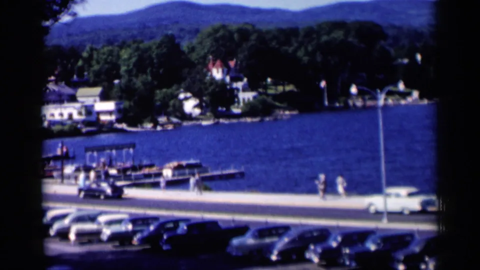 Lake George waterfront in 1955 with pedestrians, mid-century cars, marina docks, boats, shoreline homes, and wooded hills in view.