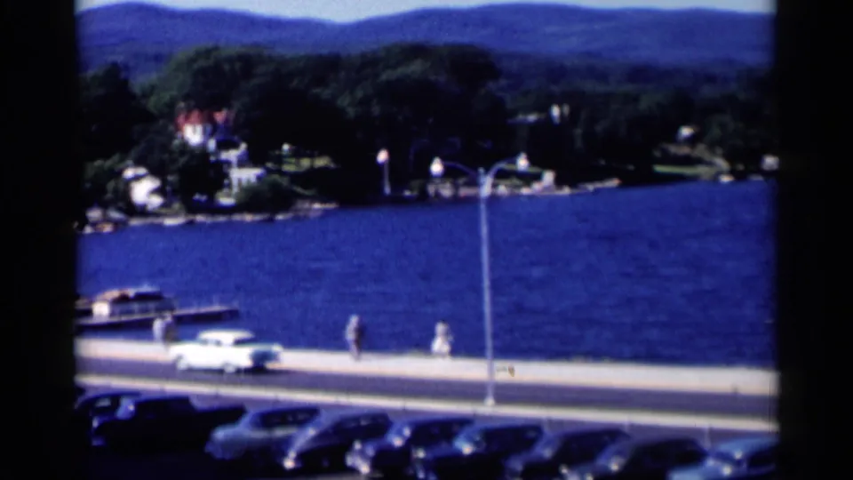 A white mid-century car travels beside Lake George while pedestrians, parked cars, docks, trees, and distant hills frame the waterfront.