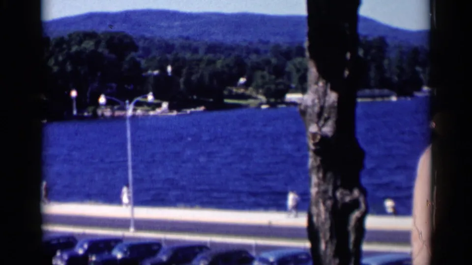 Lake George waterfront view from shade, showing pedestrians near the road, parked cars below, blue water, trees, and wooded hills.