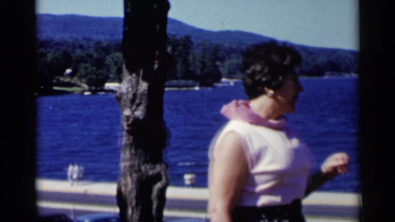 Woman in a sleeveless pink top turns beside a tree with lake water behind her.