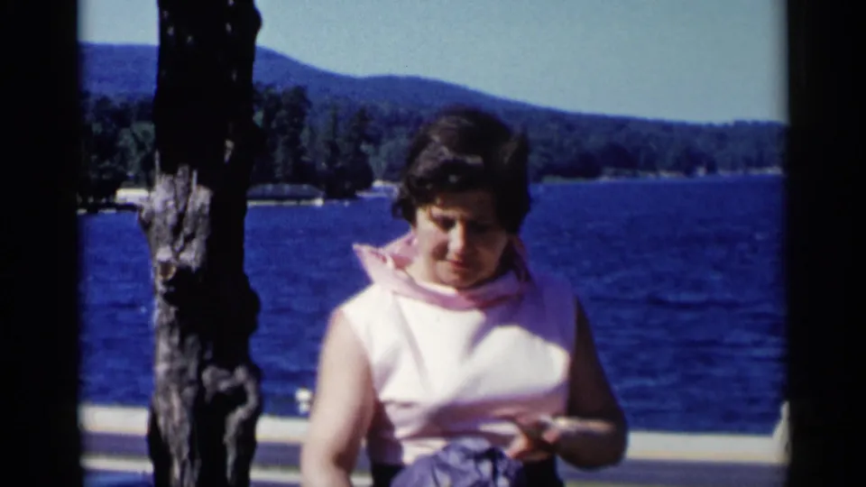 Adult woman in pink looks down near the Lake George railing, with blue water, distant wooded hills, and a paved walkway behind her.