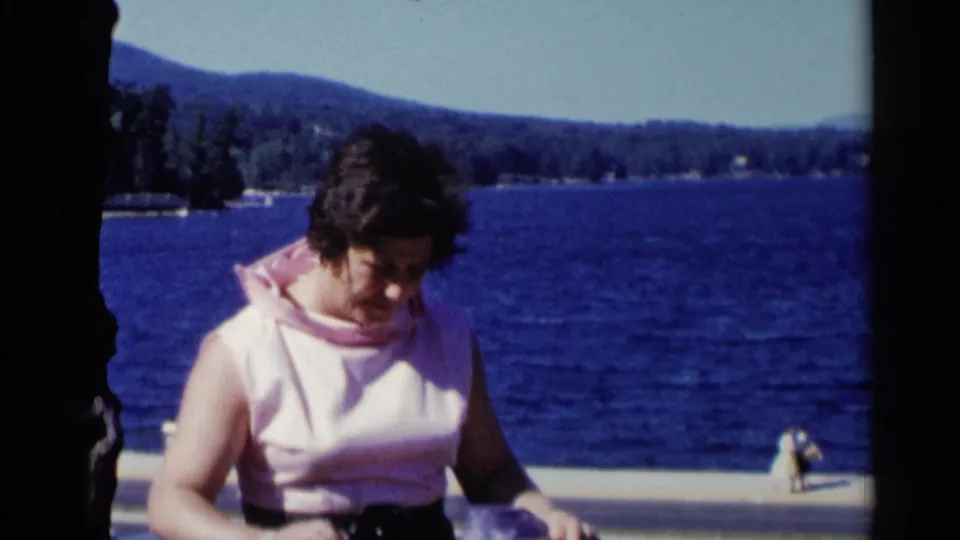 Adult woman at Lake George looks down and adjusts her purse near a tree, with deep blue water and a distant shoreline behind her.