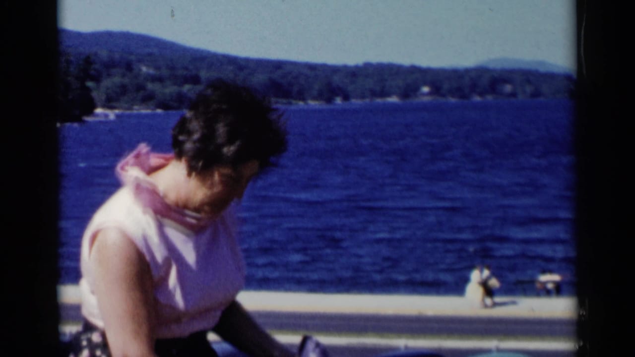 Woman in a pink sleeveless top near a lakeside road with blue water and distant hills behind her.