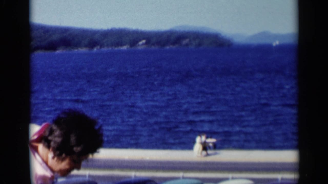 At the Lake George waterfront, the woman in pink bends partly out of frame while the lake, far shore, and small promenade figures remain visible.
