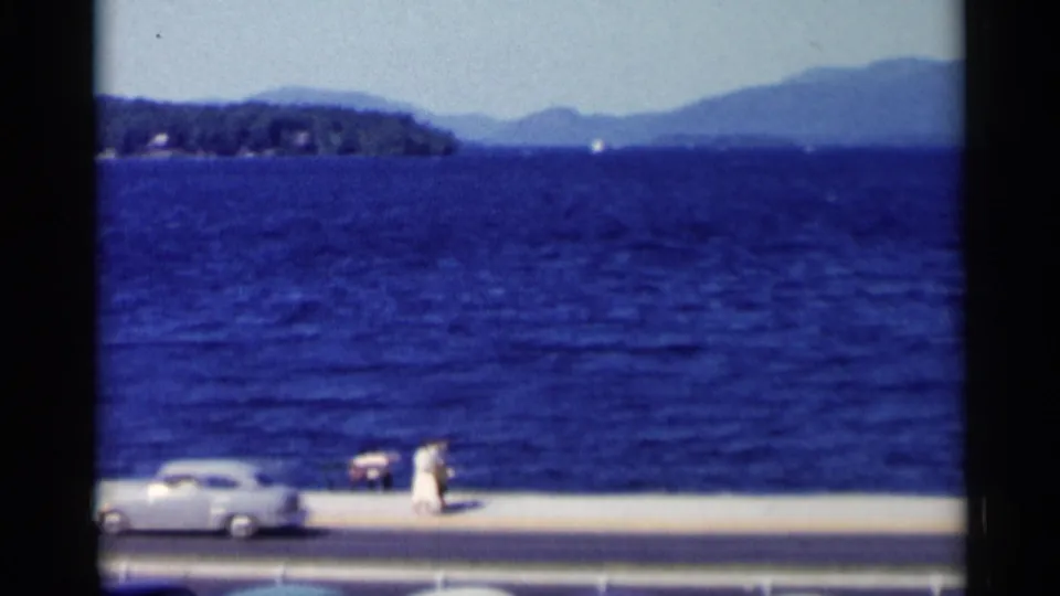 A 1955 Lake George view shows blue water and mountains above a lakeside road where a classic car passes a person in white.