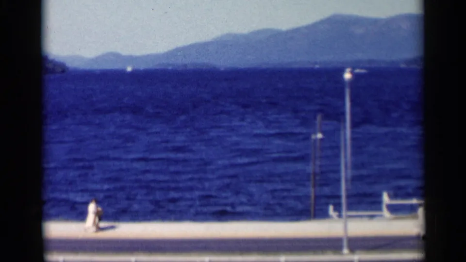 The Lake George waterfront appears with deep blue water, distant mountains, promenade poles, a person in white, and a small boat near the right edge.