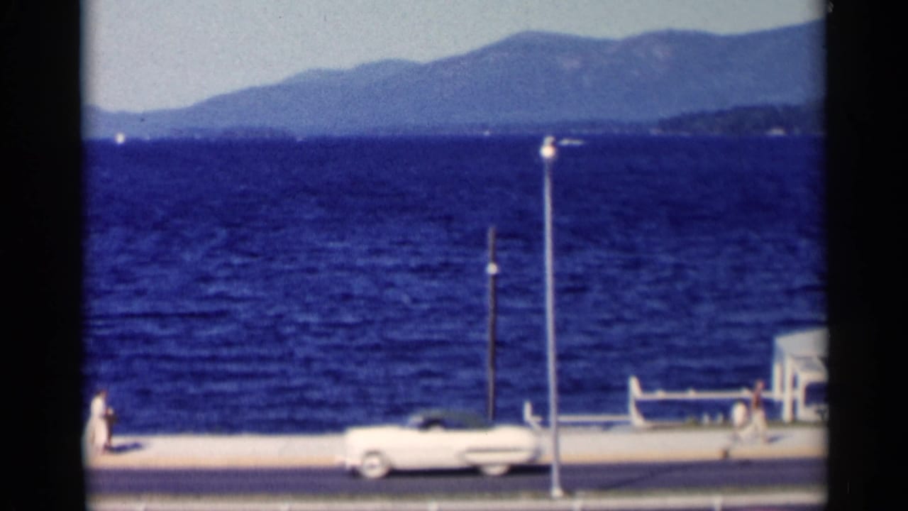 Lakeside road with a white car, promenade, blue water, and distant mountains.