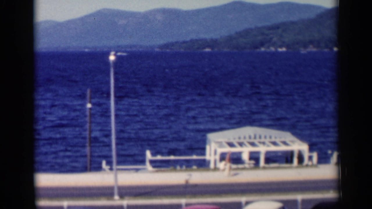 Lake George promenade with a white pavilion, benches, lampposts, blue water, and mountains beyond the shore.