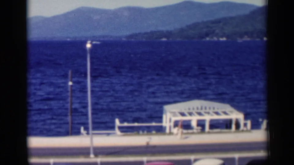 Lake George promenade with a white pavilion, benches, lampposts, blue water, and mountains beyond the shore.