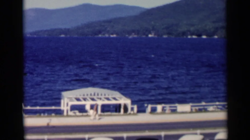 A visitor-shot view of the Lake George waterfront shows the pavilion, benches, pedestrians, blue water, and wooded hills.