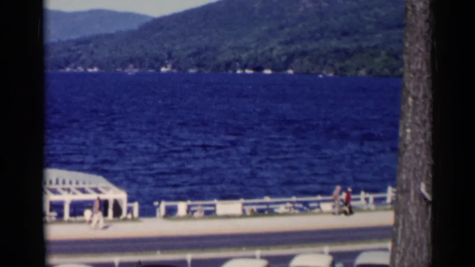 Lake George shoreline scene with pedestrians on the promenade near a white pavilion, a road in front, and mountains across the lake.