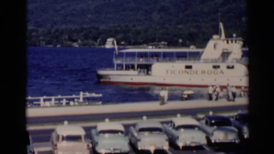 The Ticonderoga ferry is docked at Lake George in 1955, with people near the pier and classic cars parked along the waterfront.