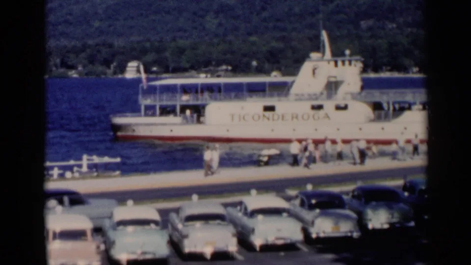 A closer 1955 Lake George view of the Ticonderoga ferry docked beside pedestrians, a white railing, and rows of classic cars.