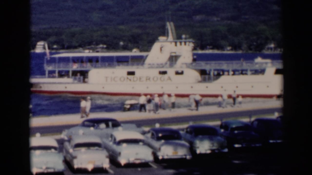 1955 Lake George home-movie frame showing the Ticonderoga ferry at a dock with parked cars and people along the waterfront.