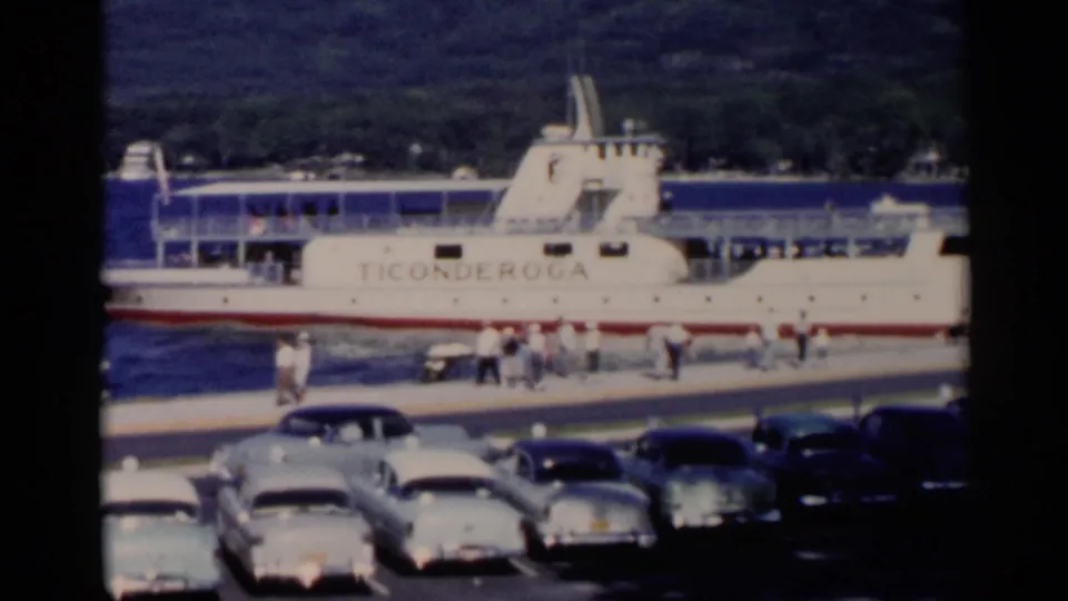 1955 Lake George home-movie frame showing the Ticonderoga ferry at a dock with parked cars and people along the waterfront.