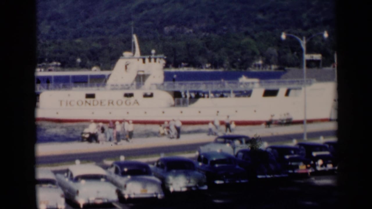 Lake George waterfront view of the Ticonderoga ferry, pier activity, parked 1950s cars, and wooded hills in the background.