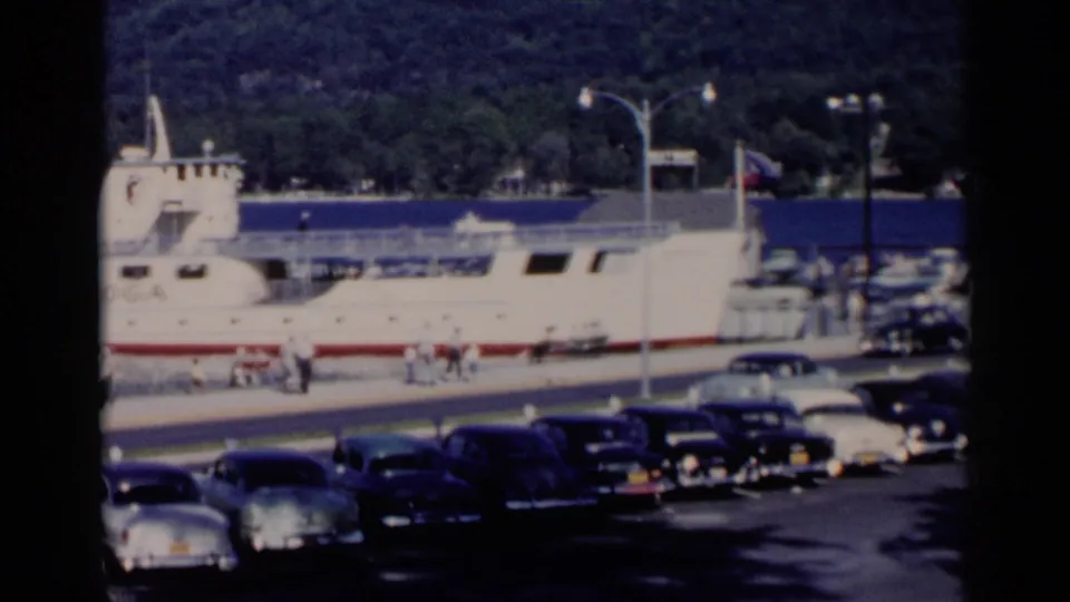 Home-movie frame of the Lake George dock area with the ferry partly in view, visitors near the pier, and rows of 1950s cars below.